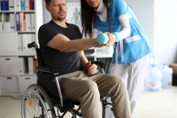 Close-up of professional physician helping patient to work out injured arm. Nurse in working uniform. Man sitting in wheelchair. Modern medicine and hospital concept