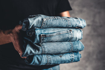 close up stack of folded denim blue jeans in hand over gray wall background, copy space