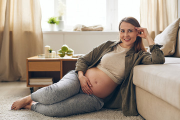 Portrait of smiling young pregnant woman sitting on floor and leaning on sofa while resting on maternity leave at home