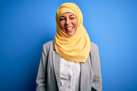 Middle age brunette business woman wearing muslim traditional hijab over blue background winking looking at the camera with sexy expression, cheerful and happy face.