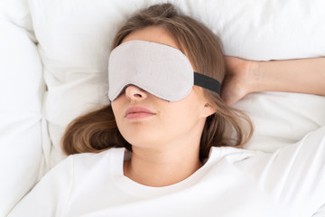Headshot of young woman dreaming in bed in daytime, wearing gray sleeping mask covering eyes fully,...