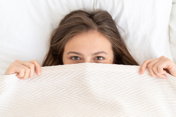Young female lying in bed in morning on pillows, covering half of face with blanket or sheet,...