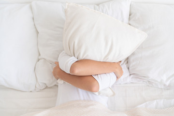 Obraz na płótnie Canvas Young girl embracing pillow, covering her face while lying in bed in white T-shirt, having fun, not ready to get up, relaxing late in morning
