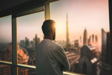 Arabic business man looking out through the office balcony seen through glass window. arab young man looking at Dubai city through hotel window. - obrazy, fototapety, plakaty