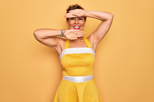 Middle Age Senior Pin Up Woman Wearing 50s Style Retro Dress Over Yellow Background Smiling Cheerful Playing Peek A Boo With Hands Showing Face. Surprised And Exited