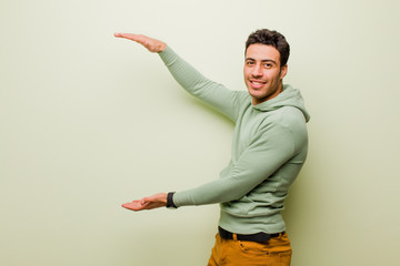 young arabian man smiling, feeling happy, positive and satisfied, holding or showing object or concept on copy space against flat wall