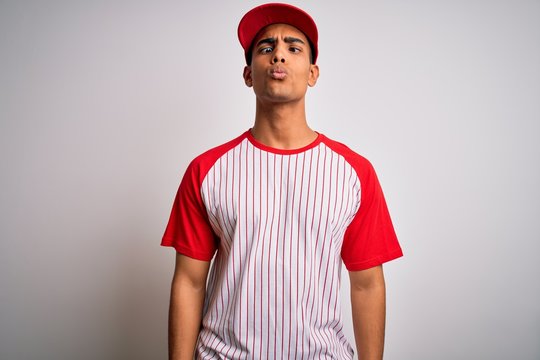 Young handsome african american sportsman wearing striped baseball t-shirt and cap making fish face with lips, crazy and comical gesture. Funny expression.