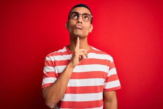 Young handsome african american man wearing casual striped t-shirt and glasses Thinking concentrated about doubt with finger on chin and looking up wondering