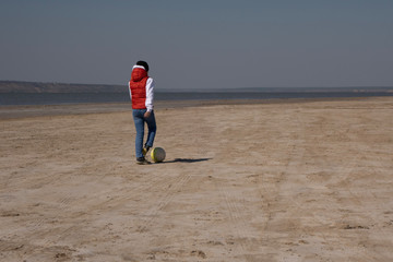 A boy of 10 years old in a white sweatshirt and orange vest plays football on a deserted beach in solitude.