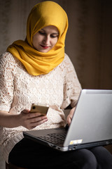 Modern Muslim working on the laptop, looking at the phone in her hand