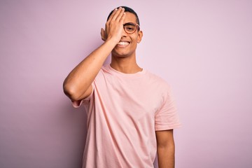 Handsome african american man wearing casual t-shirt and glasses over pink background covering one eye with hand, confident smile on face and surprise emotion.