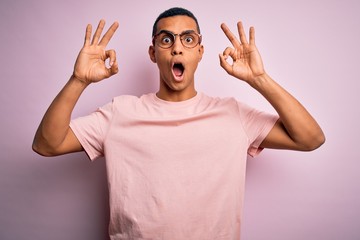 Naklejka premium Handsome african american man wearing casual t-shirt and glasses over pink background looking surprised and shocked doing ok approval symbol with fingers. Crazy expression