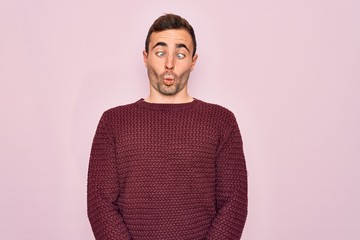 Young handsome man with blue eyes wearing casual sweater standing over pink background making fish face with lips, crazy and comical gesture. Funny expression.