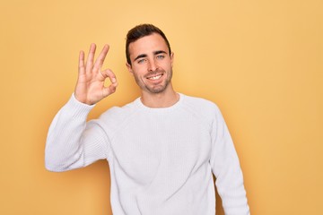 Young handsome man with blue eyes wearing casual sweater standing over yellow background smiling positive doing ok sign with hand and fingers. Successful expression.