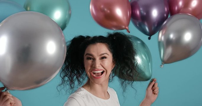 Portrait of beautiful young woman with black curly hair smiling happy with ballons in her hands in front of blue background shot in 4k super slow motion