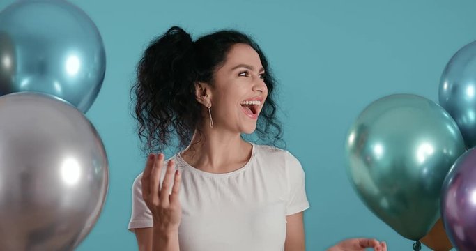 Portrait of beautiful young woman with black curly hair smiling happy surprise with ballons in her hands in front of blue background shot in 4k super slow motion