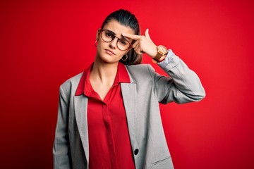 Young beautiful brunette businesswoman wearing jacket and glasses over red background pointing unhappy to pimple on forehead, ugly infection of blackhead. Acne and skin problem