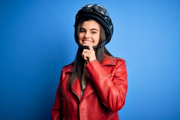 Young beautiful brunette motorcycliste woman wearing motorcycle helmet and jacket looking confident at the camera with smile with crossed arms and hand raised on chin. Thinking positive.