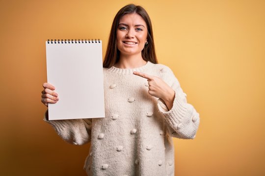 Young beautiful woman with blue eyes holding empty white notebook over yellow background very happy pointing with hand and finger