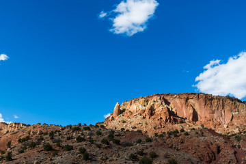 A colorful desert mesa with sandstone rock formations and cliffs under a vast blue sky illuminated by sunlight through breaks in fluffy white clouds