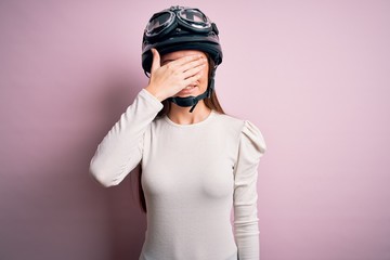 Young beautiful motorcyclist woman with blue eyes wearing moto helmet over pink background smiling and laughing with hand on face covering eyes for surprise. Blind concept.