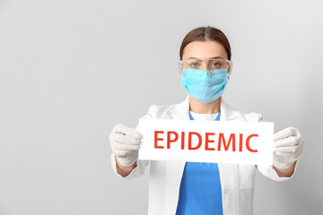 Doctor in protective mask holding paper sheet with text EPIDEMIC on grey background