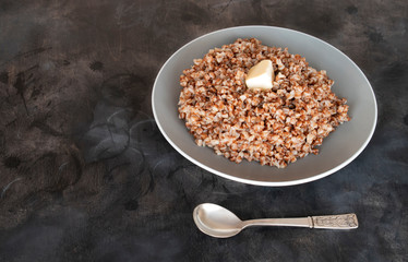 boiled buckwheat with butter in a gray plate and spoon on a dark background