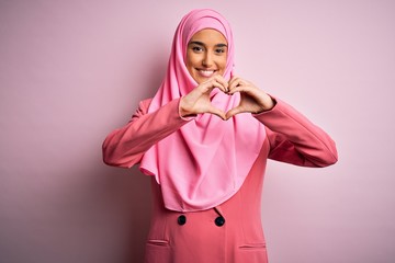 Young beautiful brunette businesswoman wearing pink muslim hijab and business jacket smiling in love doing heart symbol shape with hands. Romantic concept.