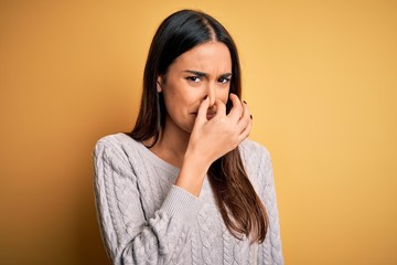 Young beautiful brunette woman wearing white casual sweater over yellow background smelling something stinky and disgusting, intolerable smell, holding breath with fingers on nose. Bad smell