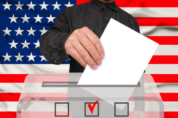 male voter lower the ballot in a transparent ballot box against the background of the national flag of the USA country, concept of state elections, referendum