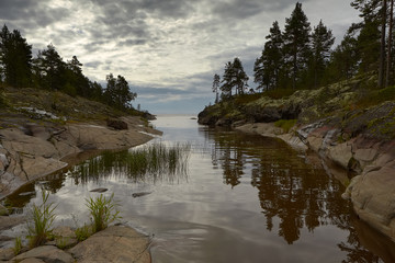 Mainly cloudy. A curving duct cuts into a stone island. You can see the exit to Lake Ladoga. Around the stones, trees, moss, lichen and heather. The water reflects the reflection of trees and sky.