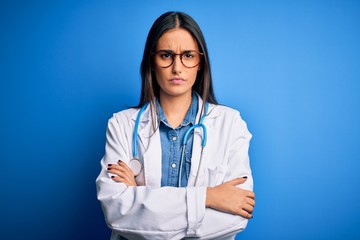 Young beautiful doctor woman wearing stethoscope and glasses over blue background skeptic and nervous, disapproving expression on face with crossed arms. Negative person.