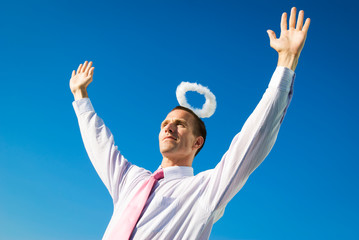 Angelic businessman standing arms raised with a halo floating above his head in the blue sky