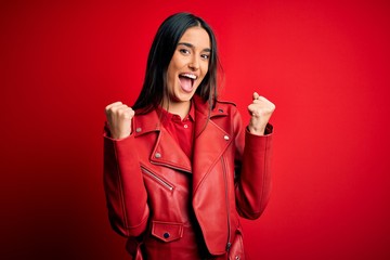 Young beautiful brunette woman wearing casual jacket standing over isolated red background celebrating surprised and amazed for success with arms raised and open eyes. Winner concept.