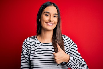 Young beautiful brunette woman wearing casual striped t-shirt over red background doing happy thumbs up gesture with hand. Approving expression looking at the camera showing success.