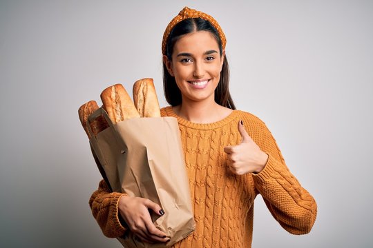 Young beautiful brunette woman holding paper bag with bread over white background happy with big smile doing ok sign, thumb up with fingers, excellent sign