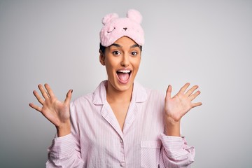 Young beautiful brunette woman wearing pajama and sleep mask over white background celebrating crazy and amazed for success with arms raised and open eyes screaming excited. Winner concept