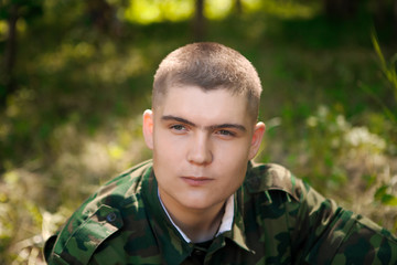 Portrait of a young soldier. A young man sits on the grass and looks ahead thoughtfully. Dressed in military uniform. In the background, green grass and shrubs.