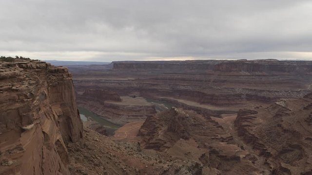 A wide timelapse looking down towards a gooseneck in the Colorado River and east towards Canyonlands National Park as seen from Dead Horse Point. A thick layer of cloud flows overhead. Vehicles on the