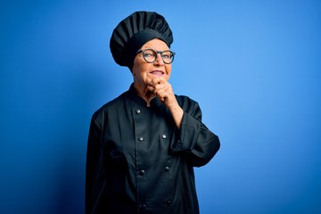 Senior beautiful grey-haired chef woman wearing cooker uniform and hat over blue background looking confident at the camera with smile with crossed arms and hand raised on chin. Thinking positive.