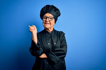 Senior beautiful grey-haired chef woman wearing cooker uniform and hat over blue background with a big smile on face, pointing with hand and finger to the side looking at the camera.