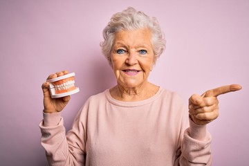 Senior beautiful grey-haired woman holding plastic denture teeth over pink background very happy pointing with hand and finger to the side