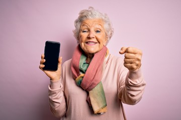 Senior beautiful grey-haired woman holding smartphone showing screen over pink background screaming proud and celebrating victory and success very excited, cheering emotion