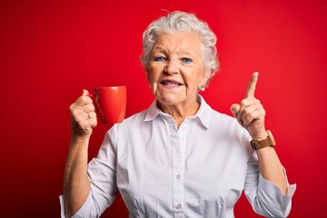 Senior beautiful woman drinking mug of coffee standing over isolated red background surprised with an idea or question pointing finger with happy face, number one