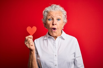 Senior beautiful woman holding paper heart standing over isolated red background scared in shock...