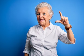 Senior beautiful woman wearing elegant shirt standing over isolated blue background smiling and confident gesturing with hand doing small size sign with fingers looking and the camera. Measure