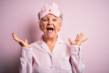 Senior beautiful woman wearing sleep mask and pajama over isolated pink background celebrating crazy and amazed for success with arms raised and open eyes screaming excited. Winner concept