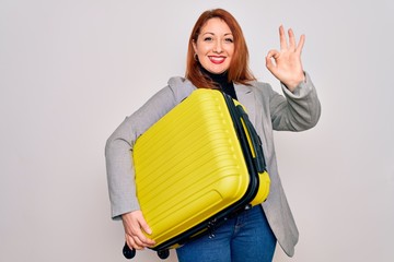 Young beautiful redhead woman holding suitcase prepared to business travel doing ok sign with fingers, excellent symbol