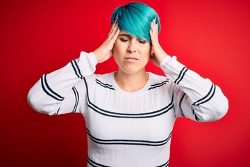 Young beautiful woman with blue fashion hair wearing striped sweater over red background suffering from headache desperate and stressed because pain and migraine. Hands on head.