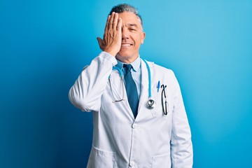 Middle age handsome grey-haired doctor man wearing coat and blue stethoscope covering one eye with hand, confident smile on face and surprise emotion.